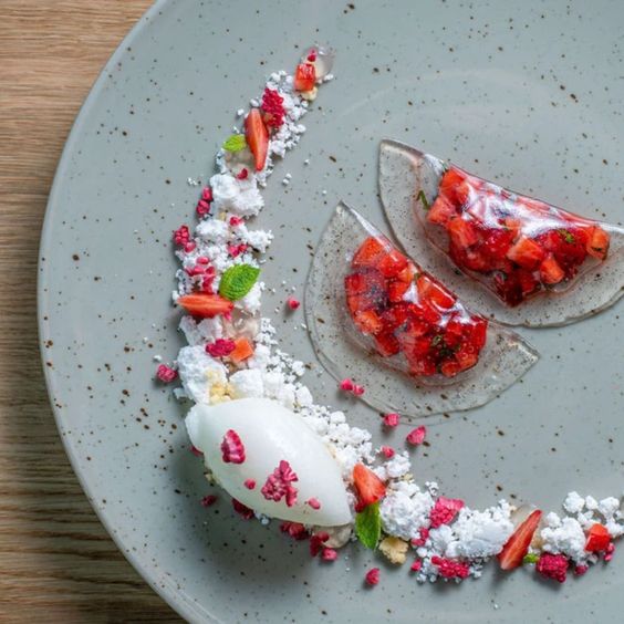 Un plato gris con unas mini empnadas transparentes de fresa, con un polvo blanco colocado en forma de media luna al lado de las empanadas de fresa.