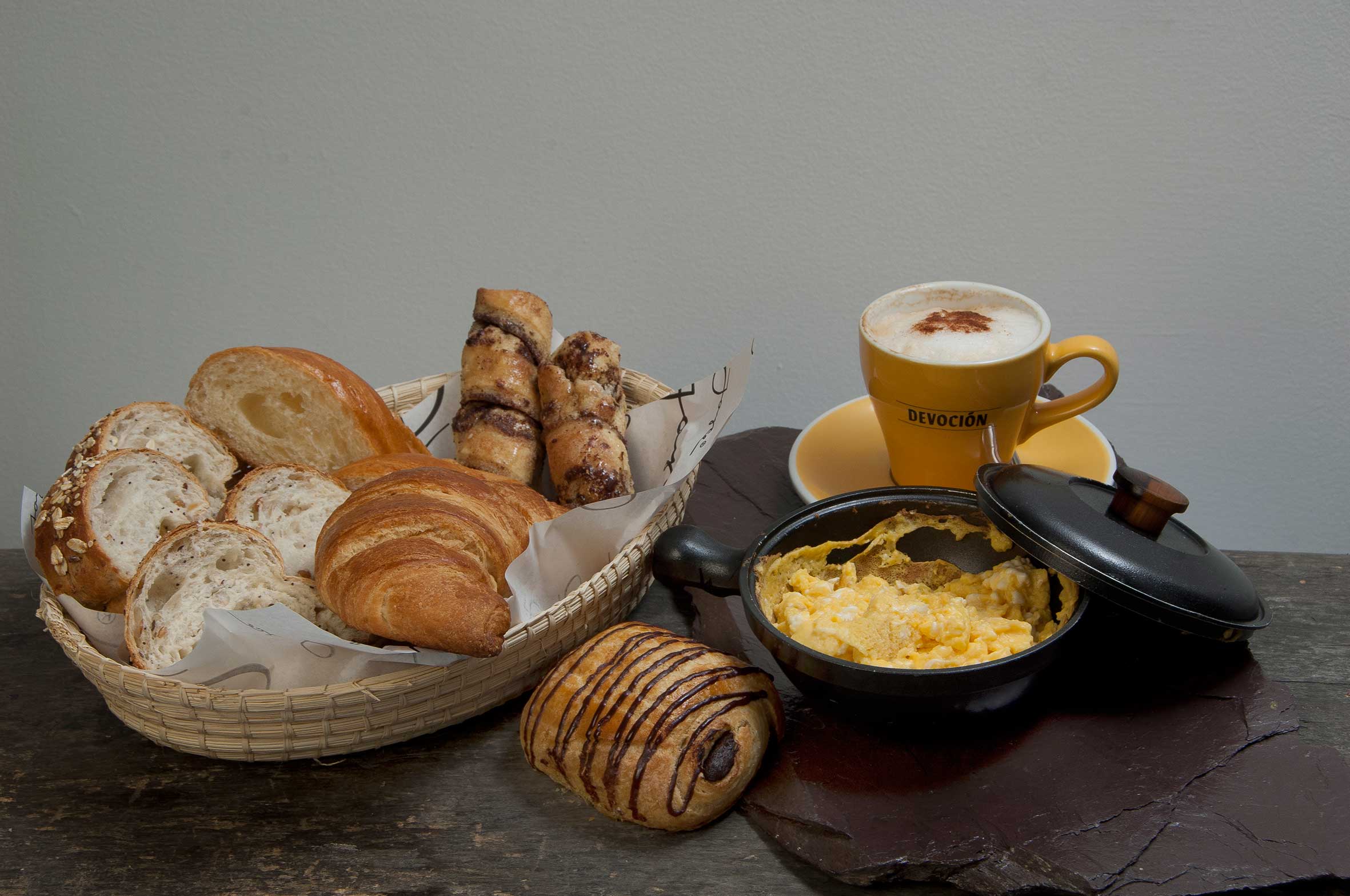 una canasta con variedad de panes, sarten con huevos revueltos, un croissant de chocolate y una taza de cafe.